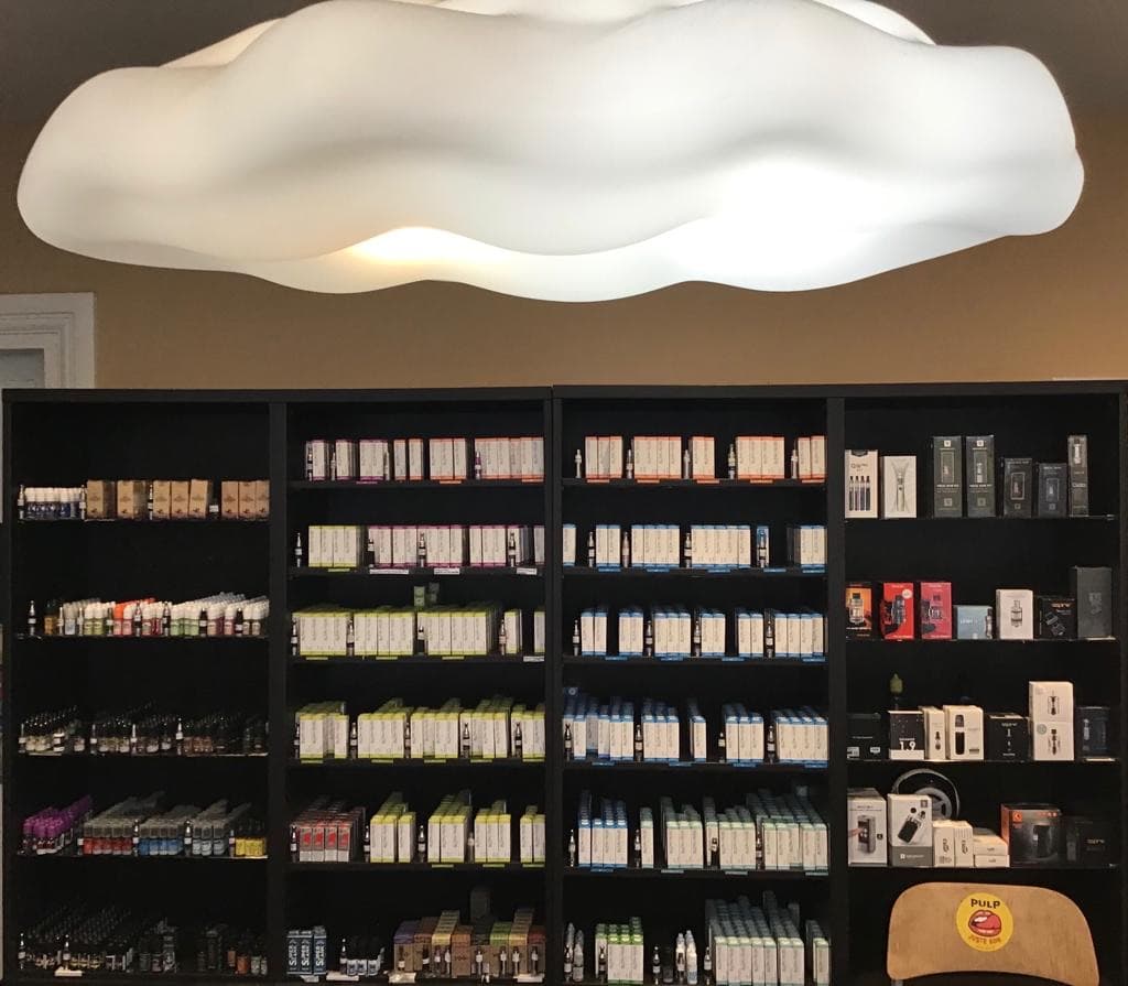 L’intérieur de la boutique : étagères noires du mur d’e-liquides et de matériel sous le plafonnier en forme de nuage